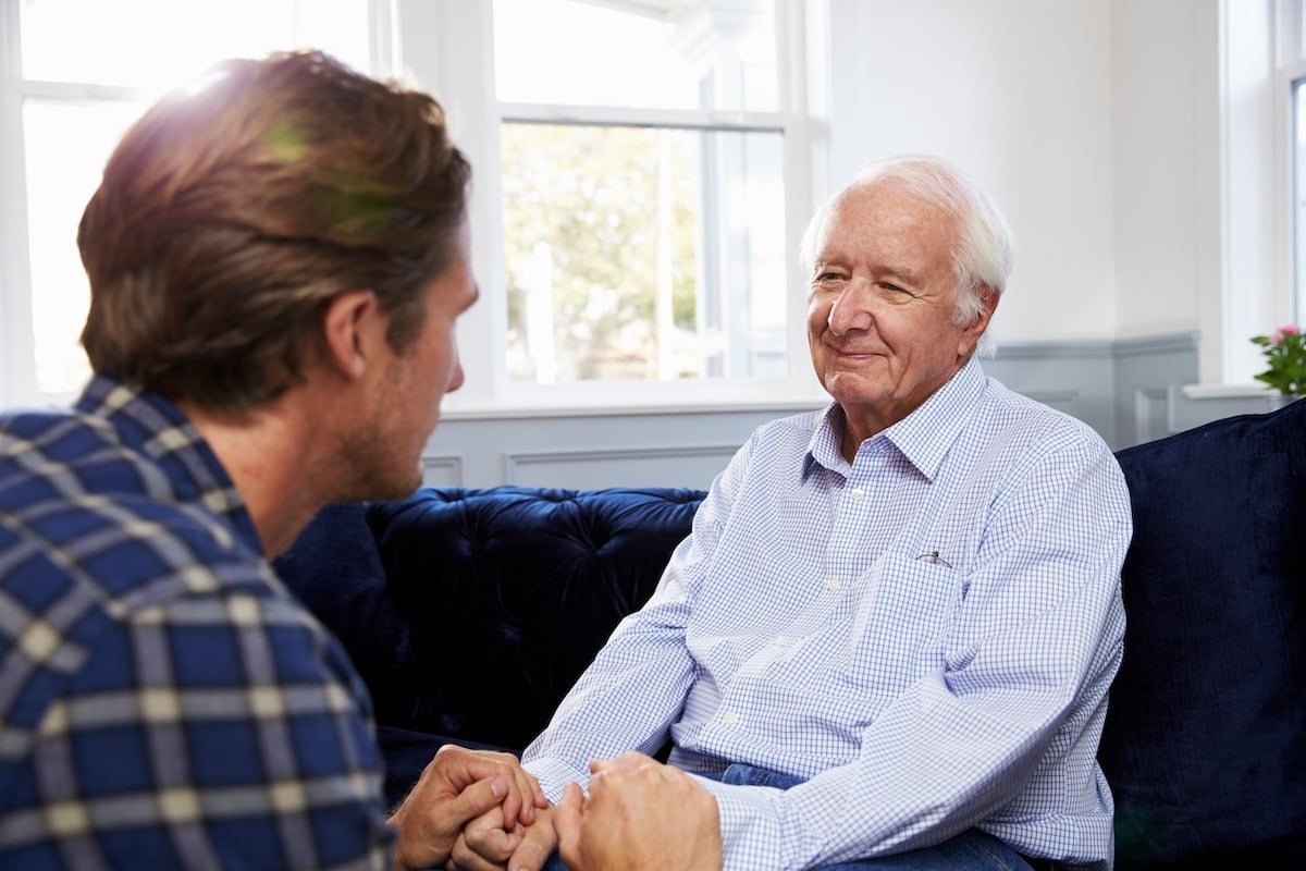 An older person smiling while holding hands with another individual on a blue sofa.