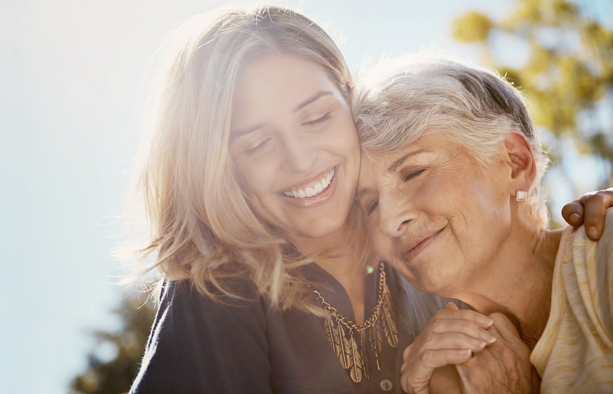 Two women, one older and one younger, smiling and embracing outdoors in sunlight.